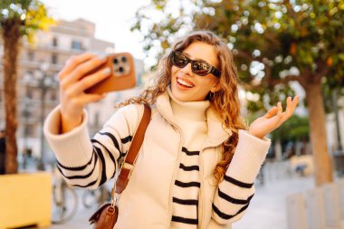 Telefonlu, güneş gözlüklü güzel bir turist gün batımında selfie çekiyor. Genç bir kadın dışarıda yürümekten hoşlanıyor. Blogculuk, turizm, eğlence kavramı.