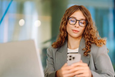 Kıvırcık saçlı ve gözlüklü genç bir kadın dışarıda dizüstü bilgisayarda çalışıyor. Kafede havanın tadını çıkaran telefonu olan güzel bir serbest çalışan kadın. İş konsepti, uzak çalışma.