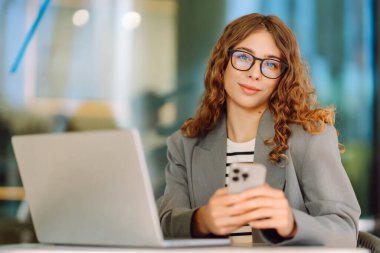 Kıvırcık saçlı ve gözlüklü genç bir kadın dışarıda dizüstü bilgisayarda çalışıyor. Kafede havanın tadını çıkaran telefonu olan güzel bir serbest çalışan kadın. İş konsepti, uzak çalışma.