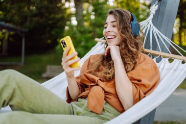 Gölü kenarındaki hamakta kablosuz kulaklık takan ve telefon kullanan güzel bir kadın. Parkta güneşli havanın tadını çıkaran genç bir kadın. Rahatlama, eğlence, blog yazma kavramı.