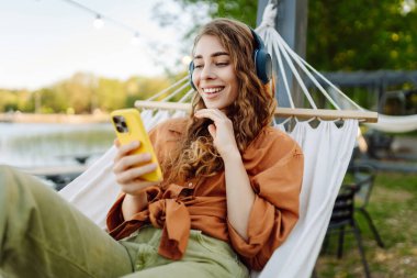 Gölü kenarındaki hamakta kablosuz kulaklık takan ve telefon kullanan güzel bir kadın. Parkta güneşli havanın tadını çıkaran genç bir kadın. Rahatlama, eğlence, blog yazma kavramı.