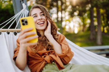 Gölü kenarındaki hamakta kablosuz kulaklık takan ve telefon kullanan güzel bir kadın. Parkta güneşli havanın tadını çıkaran genç bir kadın. Rahatlama, eğlence, blog yazma kavramı.