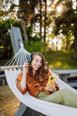Gölü kenarındaki hamakta kablosuz kulaklık takan ve telefon kullanan güzel bir kadın. Parkta güneşli havanın tadını çıkaran genç bir kadın. Rahatlama, eğlence, blog yazma kavramı.