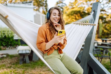 Gölü kenarındaki hamakta kablosuz kulaklık takan ve telefon kullanan güzel bir kadın. Parkta güneşli havanın tadını çıkaran genç bir kadın. Rahatlama, eğlence, blog yazma kavramı.