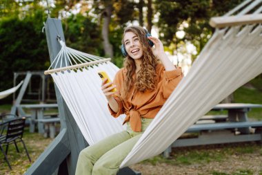 Gölü kenarındaki hamakta kablosuz kulaklık takan ve telefon kullanan güzel bir kadın. Parkta güneşli havanın tadını çıkaran genç bir kadın. Rahatlama, eğlence, blog yazma kavramı.