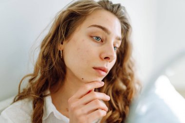 Genç bir kadının portresi küçük bir aynada yansımasına bakıyor, saçını düzeltiyor. Güzel bir kadın tenine dokunur, sivilcelerini inceler. Cilt bakımı, güzellik, doğallık kavramı.