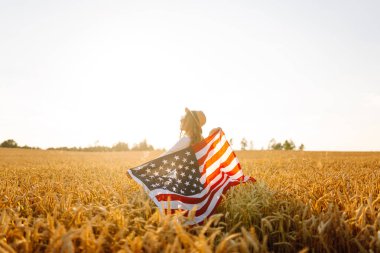 Hasır şapkalı mutlu kadının günbatımı tarlasında Amerikan bayrağını tutuşu. Genç bir kadın dışarıda eğleniyor ve özgürlüğü hissediyor. Tatil, eğlenceli konsept.