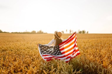 Hasır şapkalı mutlu kadının günbatımı tarlasında Amerikan bayrağını tutuşu. Genç bir kadın dışarıda eğleniyor ve özgürlüğü hissediyor. Tatil, eğlenceli konsept.