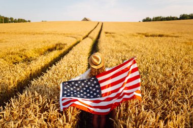 Hasır şapkalı mutlu kadının günbatımı tarlasında Amerikan bayrağını tutuşu. Genç bir kadın dışarıda eğleniyor ve özgürlüğü hissediyor. Tatil, eğlenceli konsept.