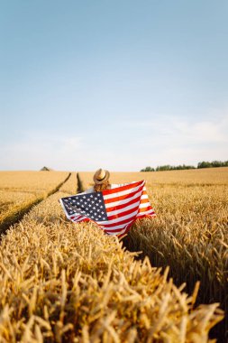 Hasır şapkalı mutlu kadının günbatımı tarlasında Amerikan bayrağını tutuşu. Genç bir kadın dışarıda eğleniyor ve özgürlüğü hissediyor. Tatil, eğlenceli konsept.