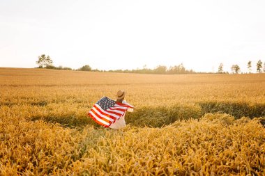 Hasır şapkalı mutlu kadının günbatımı tarlasında Amerikan bayrağını tutuşu. Genç bir kadın dışarıda eğleniyor ve özgürlüğü hissediyor. Tatil, eğlenceli konsept.