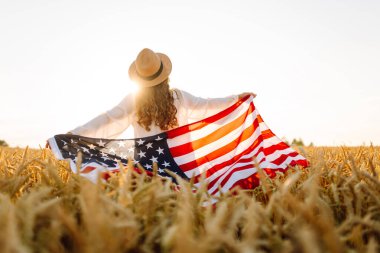 Hasır şapkalı mutlu kadının günbatımı tarlasında Amerikan bayrağını tutuşu. Genç bir kadın dışarıda eğleniyor ve özgürlüğü hissediyor. Tatil, eğlenceli konsept.