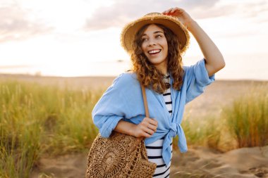 Hasır şapkalı gülümseyen kadın güneşli bir sahilde eğleniyor. Güzel bir kadın hafta sonunun tadını çıkarıyor ve dışarıda vakit geçiriyor. Tatil, keyif konsepti.