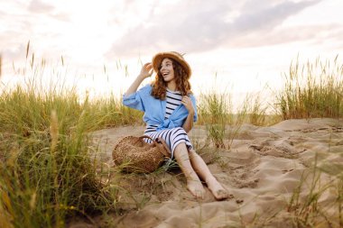 Hasır şapkalı gülümseyen kadın güneşli bir sahilde eğleniyor. Güzel bir kadın hafta sonunun tadını çıkarıyor ve dışarıda vakit geçiriyor. Tatil, keyif konsepti.