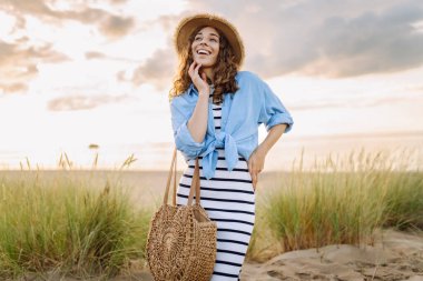 Hasır şapkalı gülümseyen kadın güneşli bir sahilde eğleniyor. Güzel bir kadın hafta sonunun tadını çıkarıyor ve dışarıda vakit geçiriyor. Tatil, keyif konsepti.