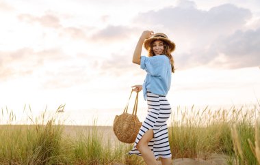 Hasır şapkalı gülümseyen kadın güneşli bir sahilde eğleniyor. Güzel bir kadın hafta sonunun tadını çıkarıyor ve dışarıda vakit geçiriyor. Tatil, keyif konsepti.