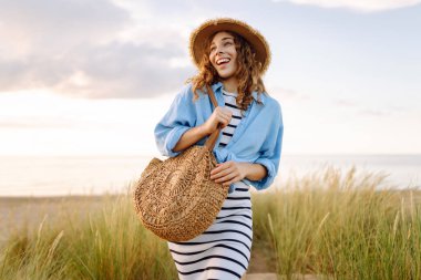 Hasır şapkalı gülümseyen kadın güneşli bir sahilde eğleniyor. Güzel bir kadın hafta sonunun tadını çıkarıyor ve dışarıda vakit geçiriyor. Tatil, keyif konsepti.