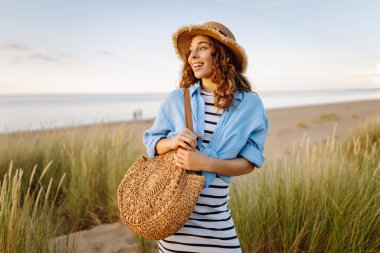 Hasır şapkalı gülümseyen kadın güneşli bir sahilde eğleniyor. Güzel bir kadın hafta sonunun tadını çıkarıyor ve dışarıda vakit geçiriyor. Tatil, keyif konsepti.