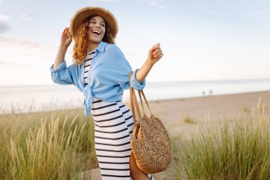 Hasır şapkalı gülümseyen kadın güneşli bir sahilde eğleniyor. Güzel bir kadın hafta sonunun tadını çıkarıyor ve dışarıda vakit geçiriyor. Tatil, keyif konsepti.