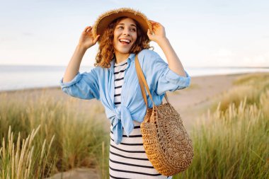 Hasır şapkalı gülümseyen kadın güneşli bir sahilde eğleniyor. Güzel bir kadın hafta sonunun tadını çıkarıyor ve dışarıda vakit geçiriyor. Tatil, keyif konsepti.