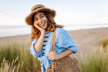 Hasır şapkalı gülümseyen kadın güneşli bir sahilde eğleniyor. Güzel bir kadın hafta sonunun tadını çıkarıyor ve dışarıda vakit geçiriyor. Tatil, keyif konsepti.