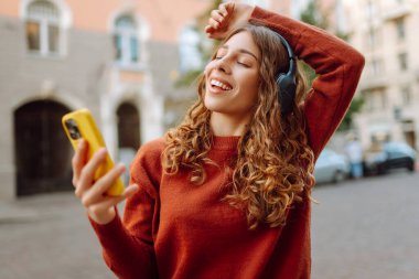 Kablosuz kulaklıklı ve telefonlu hoş bir kadın şehrin gün batımı caddelerinde yürümekten zevk alıyor. Dışarıda kulaklıkla müzik dinleyen güzel bir turist. Rahatlama konsepti.