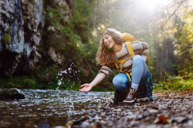 Sırtında sarı bir sırt çantasıyla gülümseyen kadın yürüyüşçü akan dağ nehrine dokunuyor. Doğanın ve maceranın tadını çıkaran genç bir kadın. Yürüyüş, seyahat ve doğa konsepti.