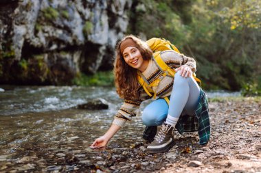 Sırtında sarı bir sırt çantasıyla gülümseyen kadın yürüyüşçü akan dağ nehrine dokunuyor. Doğanın ve maceranın tadını çıkaran genç bir kadın. Yürüyüş, seyahat ve doğa konsepti.