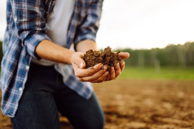 Tarım tarlasında verimli toprak taşıyan kareli gömlekli genç bir tarım uzmanının ellerine yakın çekim. Toprağın kalitesini kontrol eden bir çiftçinin elindeki kara toprak.. 