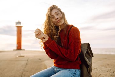 Modern elbiseli ve sırt çantalı bir kadın deniz fenerinin yanındaki iskelede eğleniyor. Güzel bir kadın deniz kenarında serin bir esintinin tadını çıkarıyor ve özgür hissediyor. Özgürlük ve rahatlama kavramı.