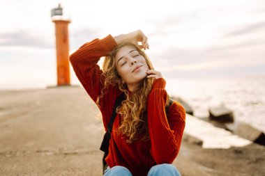 Modern elbiseli ve sırt çantalı bir kadın deniz fenerinin yanındaki iskelede eğleniyor. Güzel bir kadın deniz kenarında serin bir esintinin tadını çıkarıyor ve özgür hissediyor. Özgürlük ve rahatlama kavramı.