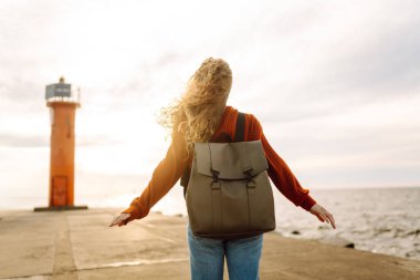 Modern elbiseli ve sırt çantalı bir kadın deniz fenerinin yanındaki iskelede eğleniyor. Güzel bir kadın deniz kenarında serin bir esintinin tadını çıkarıyor ve özgür hissediyor. Özgürlük ve rahatlama kavramı.