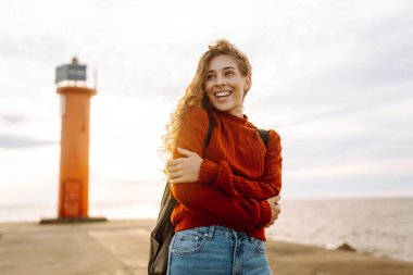 Modern elbiseli ve sırt çantalı bir kadın deniz fenerinin yanındaki iskelede eğleniyor. Güzel bir kadın deniz kenarında serin bir esintinin tadını çıkarıyor ve özgür hissediyor. Özgürlük ve rahatlama kavramı.