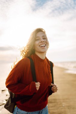 Modern elbiseli ve sırt çantalı bir kadın deniz fenerinin yanındaki iskelede eğleniyor. Güzel bir kadın deniz kenarında serin bir esintinin tadını çıkarıyor ve özgür hissediyor. Özgürlük ve rahatlama kavramı.