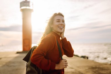Modern elbiseli ve sırt çantalı bir kadın deniz fenerinin yanındaki iskelede eğleniyor. Güzel bir kadın deniz kenarında serin bir esintinin tadını çıkarıyor ve özgür hissediyor. Özgürlük ve rahatlama kavramı.