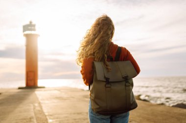Modern elbiseli ve sırt çantalı bir kadın deniz fenerinin yanındaki iskelede eğleniyor. Güzel bir kadın deniz kenarında serin bir esintinin tadını çıkarıyor ve özgür hissediyor. Özgürlük ve rahatlama kavramı.