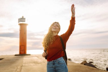 Modern elbiseli ve sırt çantalı bir kadın deniz fenerinin yanındaki iskelede eğleniyor. Güzel bir kadın deniz kenarında serin bir esintinin tadını çıkarıyor ve özgür hissediyor. Özgürlük ve rahatlama kavramı.