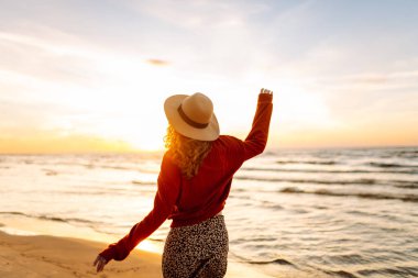 Gün batımında kumsalda yürüyen şapkalı güzel bir kadın. Genç bir kadın deniz manzarasından zevk alır ve özgür hisseder. Rahatlama, zevk kavramı..