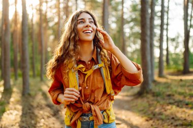 Sırt çantalı neşeli bayan gezgin gün batımında yürüyüş yapıyor. Kıvırcık saçlı güzel kadın ormanda özgürlüğü ve mutluluğu hissediyor. Macera konsepti.