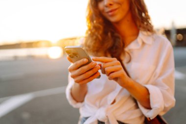 Telefonunu tutan ve günbatımının keyfini çıkaran bir kadının yakın çekimi. Güneşli bir günde setin kenarında yürüyen genç bir kadın. Bloglama, iletişim konsepti.