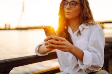 Telefonunu tutan ve günbatımının keyfini çıkaran bir kadının yakın çekimi. Güneşli bir günde setin kenarında yürüyen genç bir kadın. Bloglama, iletişim konsepti.