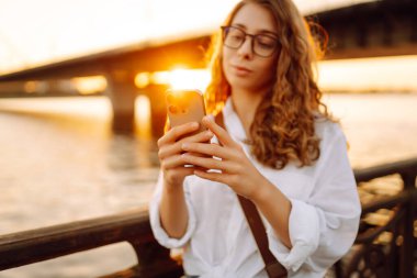 Telefonunu tutan ve günbatımının keyfini çıkaran bir kadının yakın çekimi. Güneşli bir günde setin kenarında yürüyen genç bir kadın. Bloglama, iletişim konsepti.