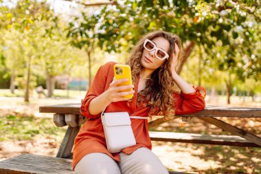 Sunny Park 'taki ahşap bankta oturan, günlük kıyafetli genç bir kadın. Güzel bir turist dışarıda dinleniyor, blog yazıyor. Boş zaman, blog konsepti.