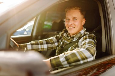Kareli tişörtle araba süren genç bir adam güneşli havanın tadını çıkarıyor. Sürüş, ulaşım, seyahat kavramı.