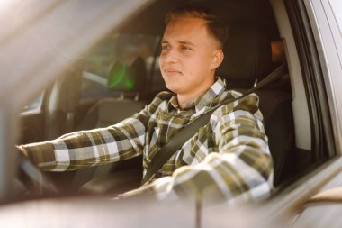 Kareli tişörtle araba süren genç bir adam güneşli havanın tadını çıkarıyor. Sürüş, ulaşım, seyahat kavramı.