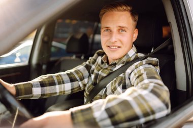 Kareli tişörtle araba süren genç bir adam güneşli havanın tadını çıkarıyor. Sürüş, ulaşım, seyahat kavramı.