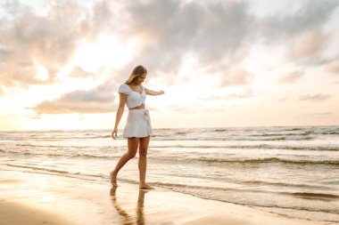 Gün batımında kumlu sahilde yürüyen mutlu genç bir kadın. Gülümseyen kadın havanın tadını çıkarıyor ve dışarıda özgürlüğü hissediyor. Tatil, hafta sonu konsepti. Yaşam biçimi.