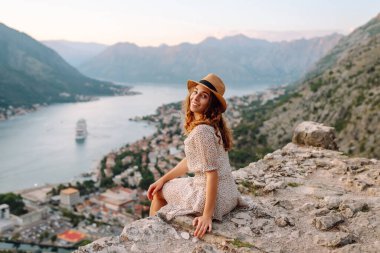 Güzel bir kadın uçurumda oturuyor ve deniz panoramik manzarasının tadını çıkarıyor. Gün batımında gözlem güvertesinde şapkalı ve elbiseli genç bir turist özgürlüğü hissediyor. Turizm konsepti.