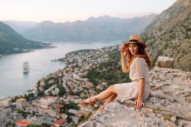 Güzel bir kadın uçurumda oturuyor ve deniz panoramik manzarasının tadını çıkarıyor. Gün batımında gözlem güvertesinde şapkalı ve elbiseli genç bir turist özgürlüğü hissediyor. Turizm konsepti.