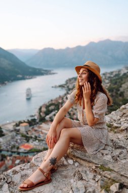 Güzel bir kadın uçurumda oturuyor ve deniz panoramik manzarasının tadını çıkarıyor. Gün batımında gözlem güvertesinde şapkalı ve elbiseli genç bir turist özgürlüğü hissediyor. Turizm konsepti.